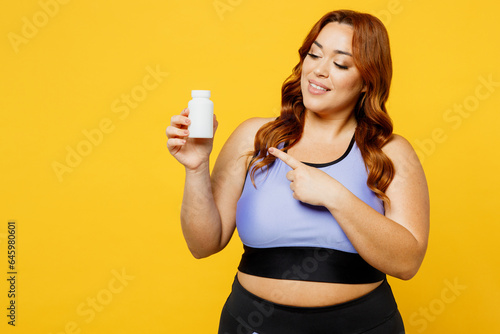 Young smiling chubby plus size big fat fit woman wear blue top warm up train point hold bottle with bio suplements vitamins isolated on plain yellow background studio home gym. Workout sport concept. © ViDi Studio