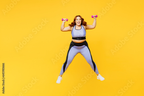 Full body excited young chubby plus size big fat fit woman wear blue top warm up train hold dumbbells jump high look camera isolated on plain yellow background studio home gym. Workout sport concept.