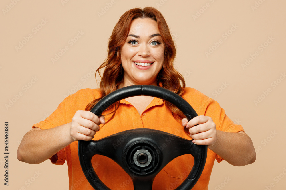 Young ginger chubby overweight woman wears orange shirt casual clothes hold steering wheel driving car look camera isolated on plain pastel light beige background studio portrait. Lifestyle concept. Young ginger chubby overweight woman wears orange shirt casual clothes hold steering wheel driving car look camera isolated on plain pastel light beige background studio portrait. Lifestyle concept.