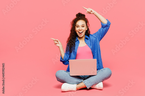 Full body young IT woman of African American ethnicity wear blue shirt casual clothes sits hold use work on laptop pc computer point aside isolated on plain pastel pink background. Lifestyle concept.