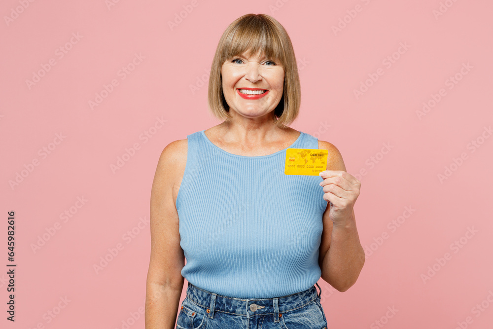 Elderly smiling cheerful blonde woman 50s years old she wears blue undershirt casual clothes hold in hand mock up of credit bank card isolated on plain pastel light pink background. Lifestyle concept. Elderly smiling cheerful blonde woman 50s years old she wears blue undershirt casual clothes hold in hand mock up of credit bank card isolated on plain pastel light pink background. Lifestyle concept.