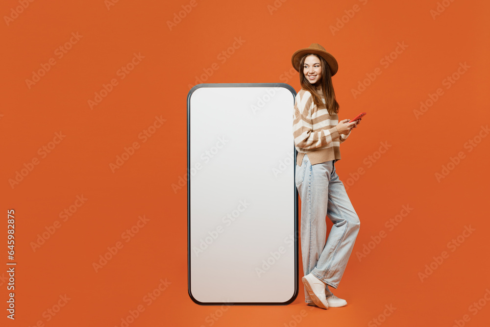 Full body side view young woman wear striped sweater hat casual clothes stand near big huge blank screen mobile cell phone with area use smartphone isolated on plain orange red color wall background.