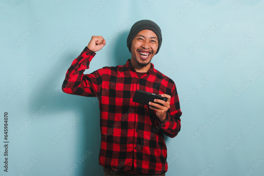 Excited young Asian man gamer with beanie hat and red plaid flannel shirt is raising his fist in a YES gesture while winning and playing an online game on his smartphone, isolated on a blue background Excited young Asian man gamer with beanie hat and red plaid flannel shirt is raising his fist in a YES gesture while winning and playing an online game on his smartphone, isolated on a blue background