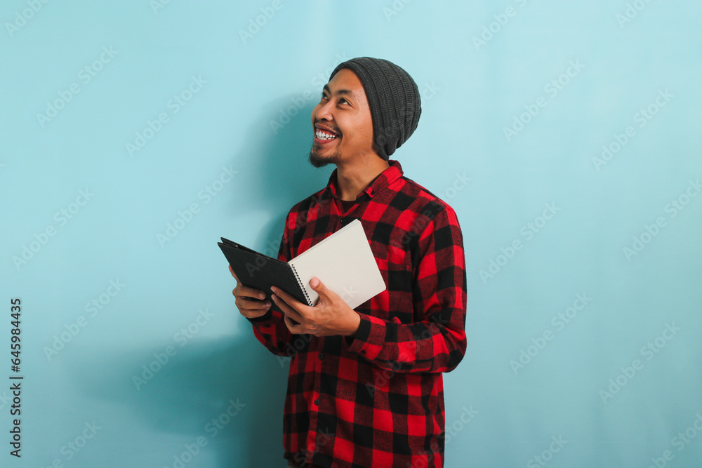 A smiling young Asian man is holding a book while standing against a blue background A smiling young Asian man is holding a book while standing against a blue background