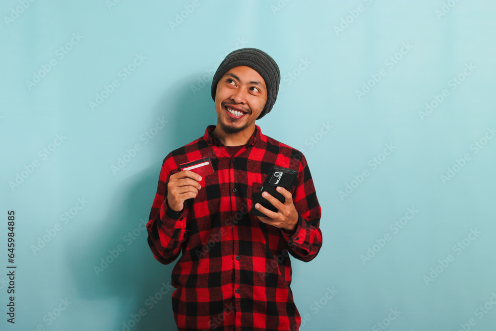Happy young Asian man is holding smartphone and banking credit card, isolated on blue background. Happy young Asian man is holding smartphone and banking credit card, isolated on blue background.