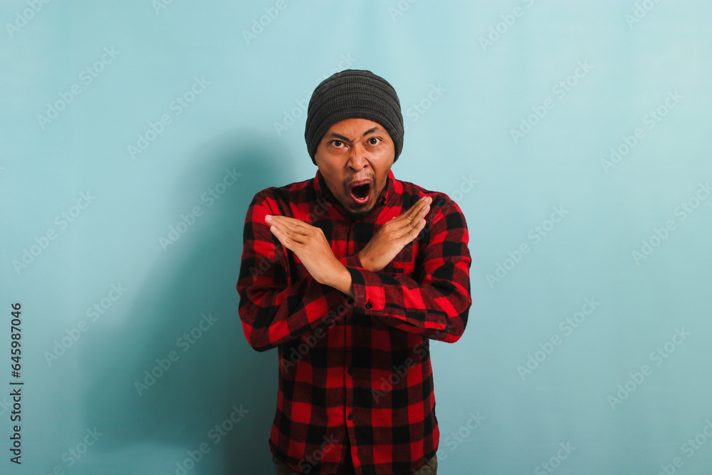 Angry young Asian man stands with his arms crossed, stop gesture, isolated on blue background. Angry young Asian man stands with his arms crossed, stop gesture, isolated on blue background.