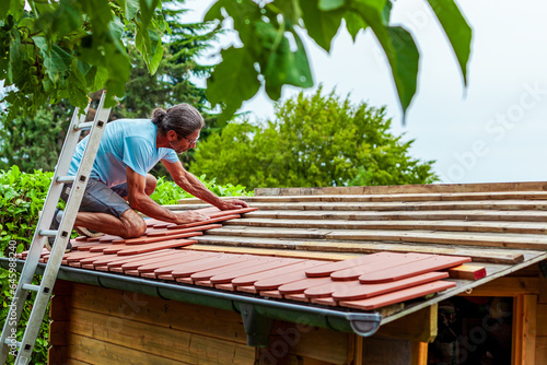 Roofer at work, installing clay roof tiles. back garden with shed, summer house garden timber outbuilding. new roof of the shed in the back yard. install tile on roof of old house.