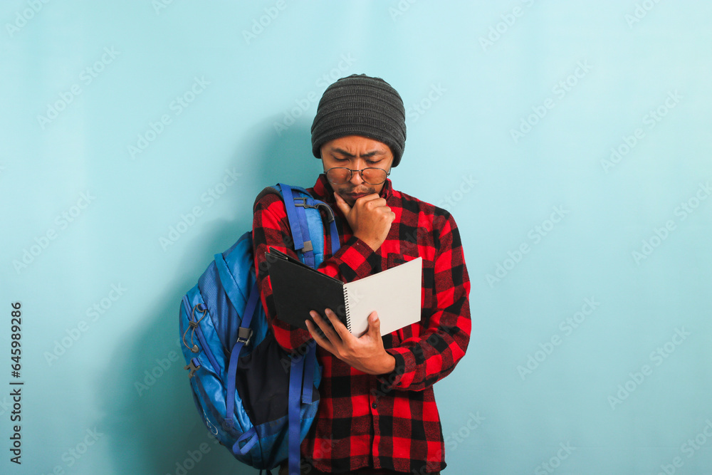 Pensive young Asian student man is thinking while holding book and standing against blue background Pensive young Asian student man is thinking while holding book and standing against blue background