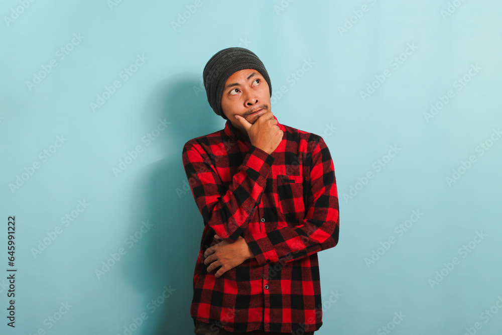 Thoughtful Young Asian man holds his chin, making plans, isolated on a blue background Thoughtful Young Asian man holds his chin, making plans, isolated on a blue background