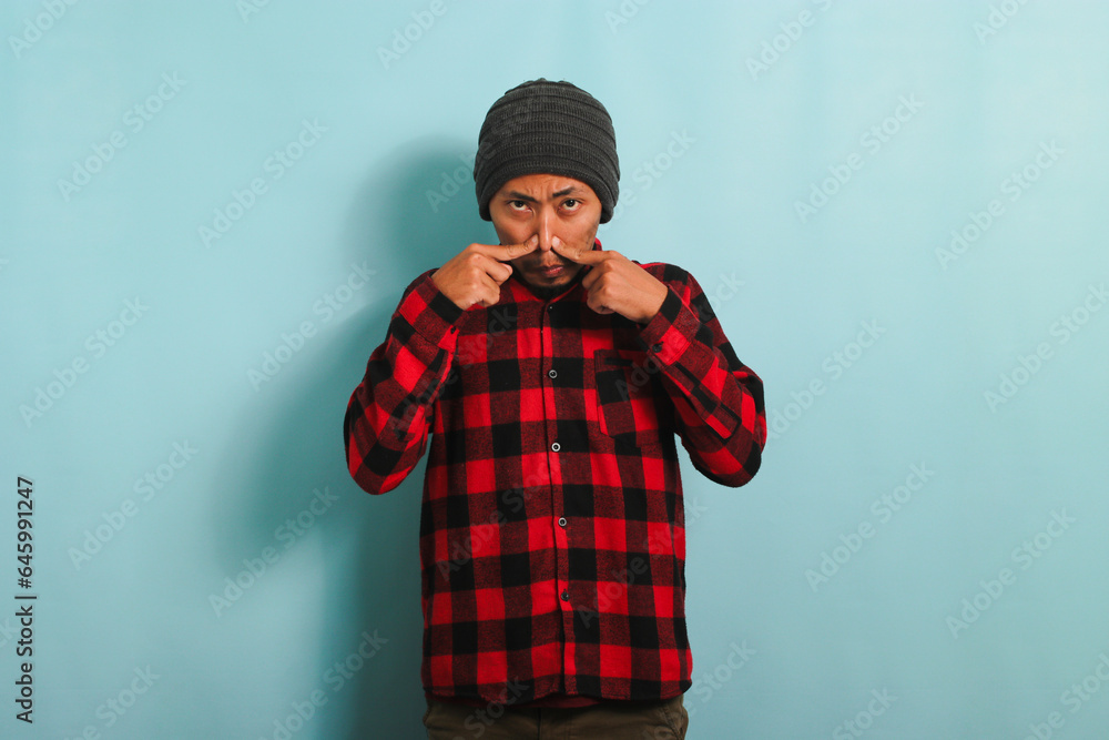 Annoyed young Asian man holds his breath smells something stinky, isolated on blue backdrop Annoyed young Asian man holds his breath smells something stinky, isolated on blue backdrop