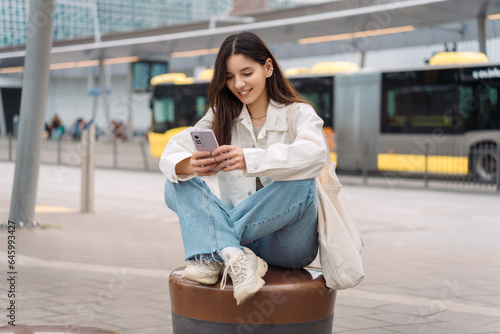 Waiting for the bus and for love. Bright cheerful young woman of Turkish mixed race in white casual clothes with a charming smile holding a phone in the street at public transportation stop