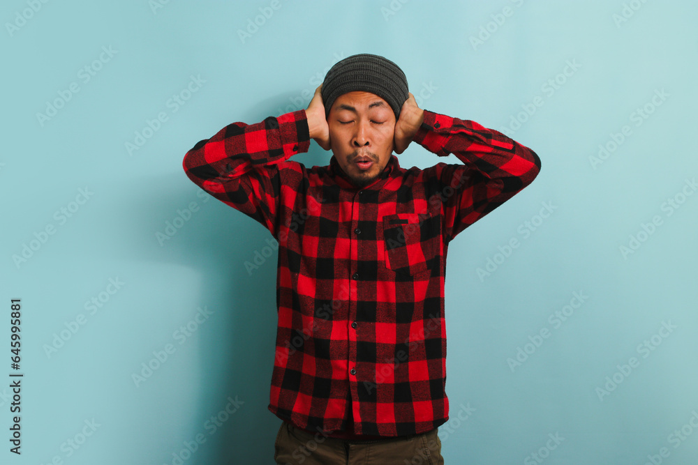 A young Asian man with a beanie hat and a red plaid flannel shirt is suffering from a headache, feeling desperate and stressed due to the pain and migraine, while standing against a blue background A young Asian man with a beanie hat and a red plaid flannel shirt is suffering from a headache, feeling desperate and stressed due to the pain and migraine, while standing against a blue background