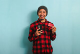 Happy young Asian man with a beanie hat and a red plaid flannel shirt is holding a smartphone and a banking credit card, doing online shopping, looking at camera, isolated on a blue background.