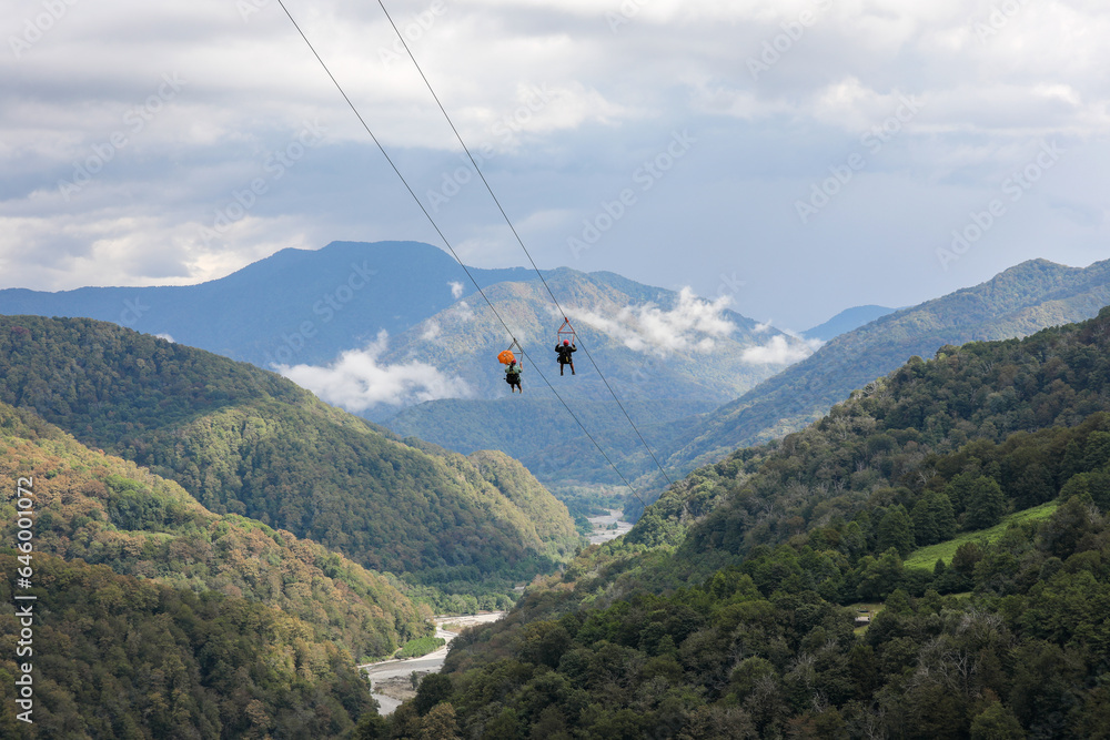 Fototapeta premium Two tourists going down on a zipline, against the backdrop of a picturesque mountain landscape. Sochi, Solokhaul, Russia.