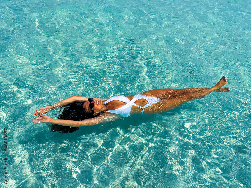 A smiling girl, adorned in white swimwear, floats gracefully on her back, embraced by the crystal-clear ocean. This snapshot embodies the carefree joy and the pristine beauty of the Maldivian seascape