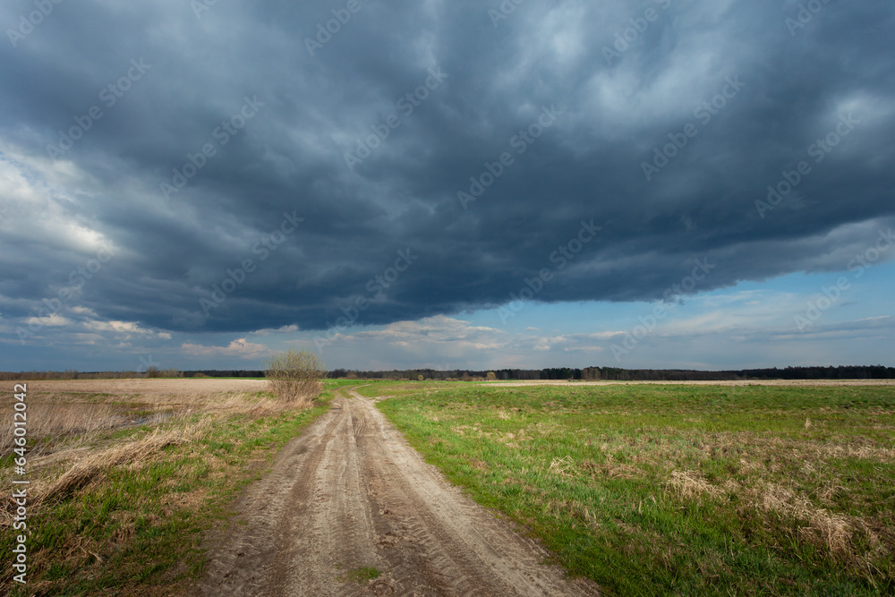 Unpaved road through meadows and dark cloud on the sky