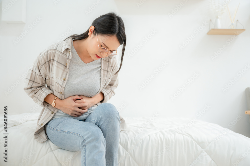 Obraz premium Portrait of Asian woman with abdominal pain sitting in bed at home She was suffering from severe abdominal pain. Stomach discomfort and food poisoning diarrhea