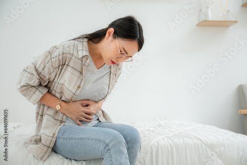  Asian woman with abdominal pain sitting in bed at home She was suffering from severe abdominal pain. Stomach discomfort and food poisoning diarrhea