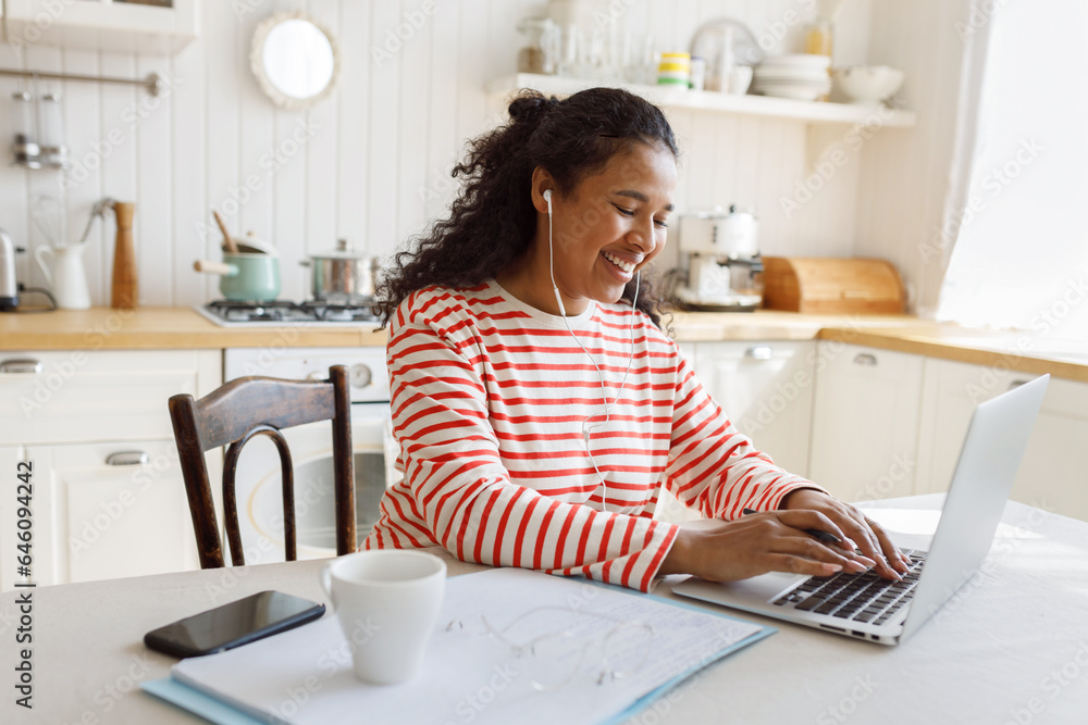 Side view of happy african american female freelancer working on laptop sitting at kitchen table, making notes in her diary, chatting online in corporate chat or messenger, wearing wired earphones Side view of happy african american female freelancer working on laptop sitting at kitchen table, making notes in her diary, chatting online in corporate chat or messenger, wearing wired earphones