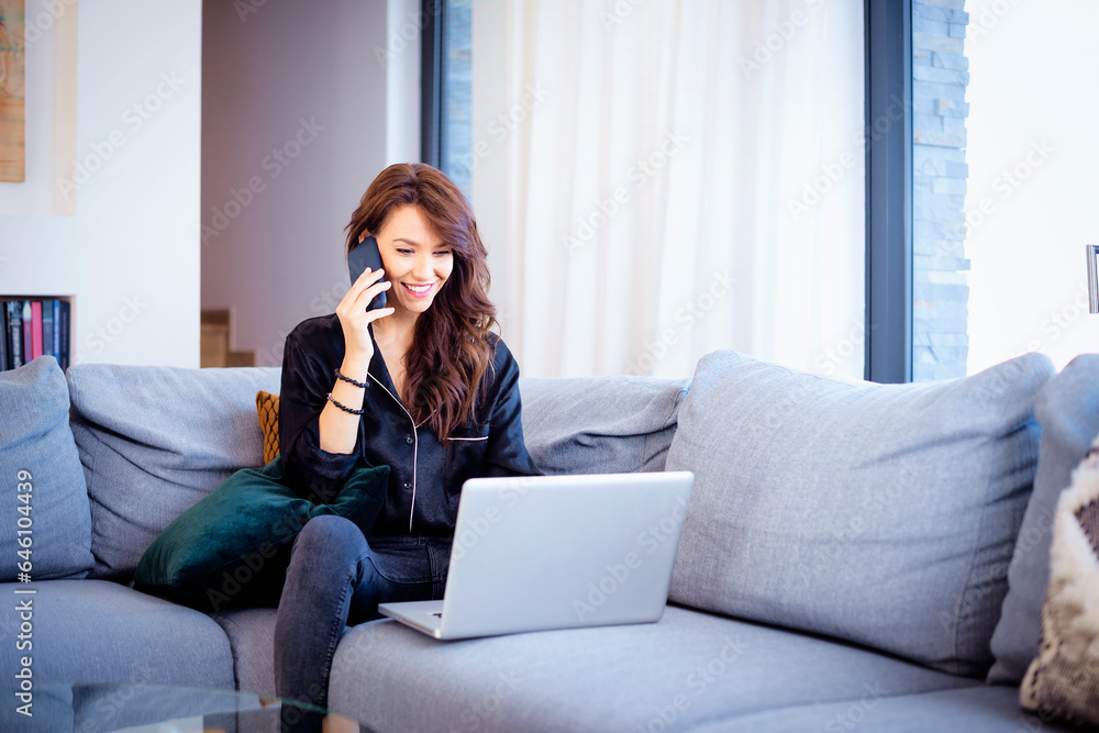 A middle-aged woman with brown hair is sitting at home using a laptop and a mobile phone A middle-aged woman with brown hair is sitting at home using a laptop and a mobile phone