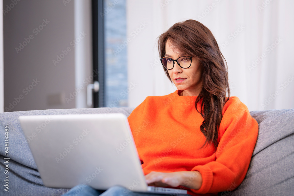 Serious faced brunette haired woman sitting on sofa at home and using laptop for work Serious faced brunette haired woman sitting on sofa at home and using laptop for work