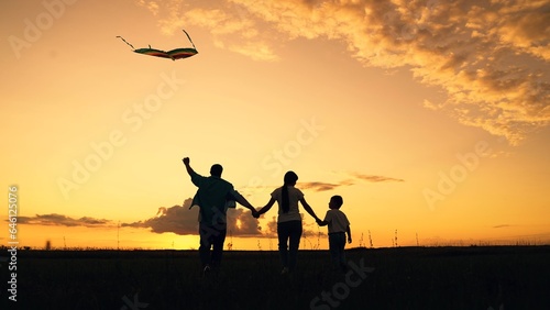 Young family is playing with kite in field. Happy family with baby in park playing with kite. Family walk on grass  child dreams of flying. People together travel  nature  silhouette. Kid wants to fly