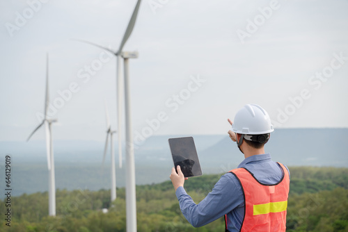A worker in a wind turbine field looks towards wind turbine while holding a tablet, managing the efficient production of renewable energy. © tope007