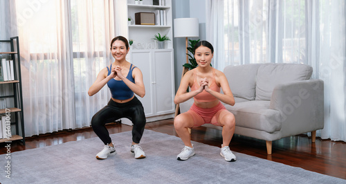 Vigorous energetic woman with trainer or workout buddy doing exercise at home. Young athletic asian woman strength and endurance training session as home workout routine with squat.