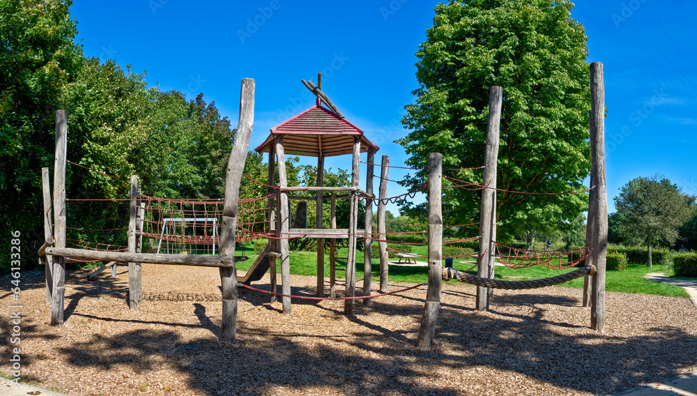 Kletterburg aus Holz auf einem menschenleeren, öffentlichen Spielplatz im Sommer bei schönem Wetter und wolkenlosem Himmel, eingesäumt von Laubbäumen Kletterburg aus Holz auf einem menschenleeren, öffentlichen Spielplatz im Sommer bei schönem Wetter und wolkenlosem Himmel, eingesäumt von Laubbäumen