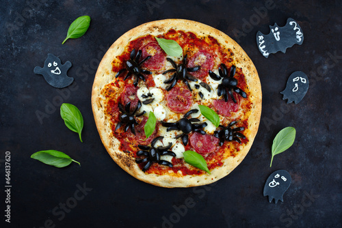 Traditionelle italienische Halloween Spinnen Pizza mit Salami, Mozzarella und Oliven serviert als Draufsicht mit Fledermaus und Geister auf einem alten rustikalen Board photo