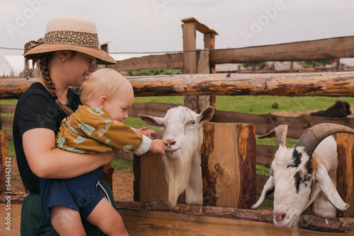 Mom and little daughter feed and care for goats on the farm. Adorable cute toddler boy with mother feeding goat on a kids farm. Beautiful baby child petting animals in the zoo. Excited and happy boy