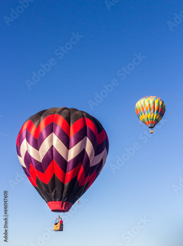 2 hot air balloons are rising in the early morning light into a blue and wispy clouded sky, 1 balloon is slightly above the balloons, The are brightly colored.