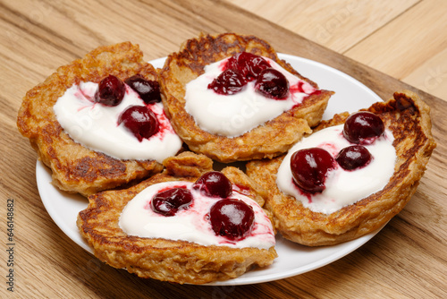 Pancakes are drizzled with creamy sour cream and sweet cherries on a white plate. 