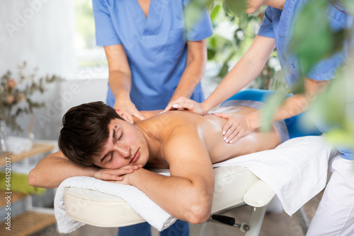 Two female masseur doing massage at back man who lying on massage table in a beauty salon