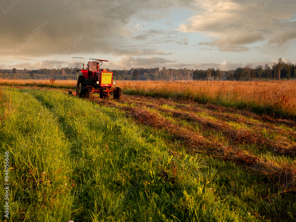 Fototapeta premium Old rusty tractor with grass cutting machine in a agriculture field with tall grass at stunning sunrise. Farming industry. Beautiful nature scene at special time. Nobody.