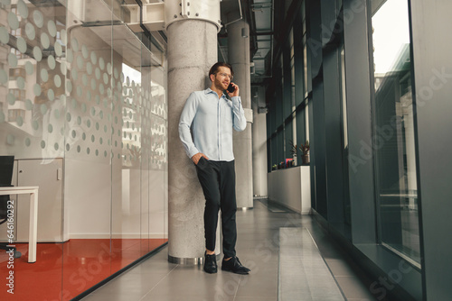 Smiling male entrepreneur talking phone with client while standing in office and looking at side 