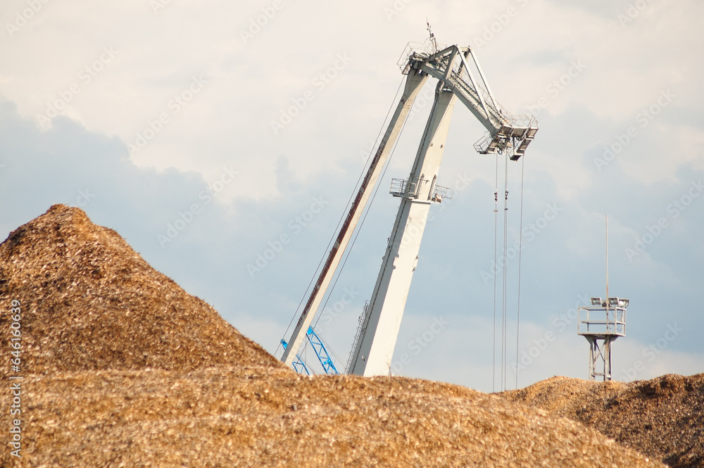 a mountain of wood chips and a cargo crane against the background of the sky and clouds