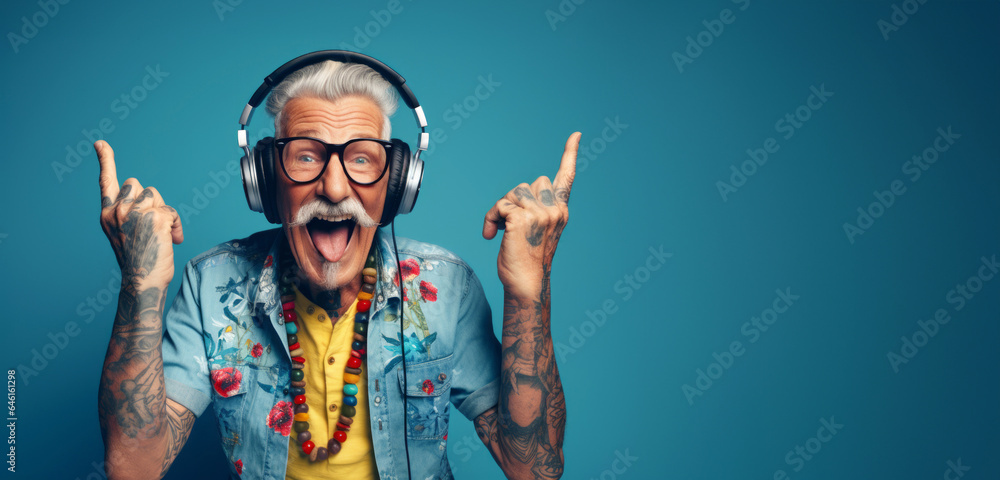 A happy hipster and cool grandfather, original style and tattoos, wearing headphones enjoying music, pointing his fingers up. Active and fun lifestyle concept for seniors: Sunset of life in colors - obrazy, fototapety, plakaty 