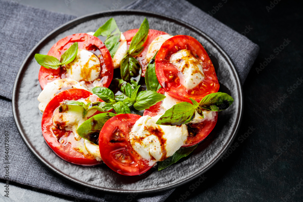 Plate of healthy classic caprese salad with ripe tomatoes and mozzarella cheese with fresh basil leaves on dark background. Close up