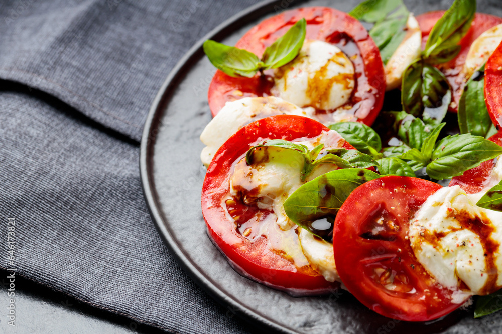 Plate of healthy classic caprese salad with ripe tomatoes and mozzarella cheese with fresh basil leaves on dark background. Close up Plate of healthy classic caprese salad with ripe tomatoes and mozzarella cheese with fresh basil leaves on dark background. Close up