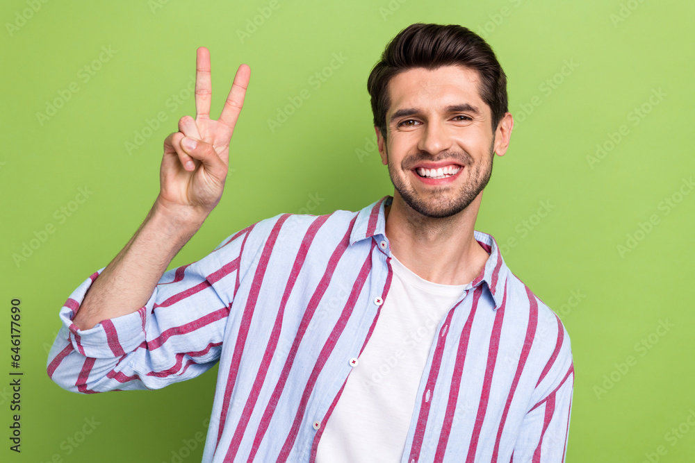 Photo of handsome nice cheerful man with stubble dressed striped shirt showing v-sign toothy smiling isolated on green color background Photo of handsome nice cheerful man with stubble dressed striped shirt showing v-sign toothy smiling isolated on green color background
