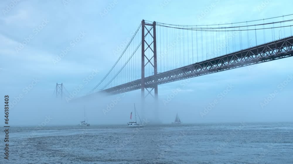 View of 25 de Abril Bridge famous tourist landmark of Lisbon connecting Lisboa and Almada in heavy fog mist with yacht boats passing under. Lisbon, Portugal, Europe View of 25 de Abril Bridge famous tourist landmark of Lisbon connecting Lisboa and Almada in heavy fog mist with yacht boats passing under. Lisbon, Portugal, Europe