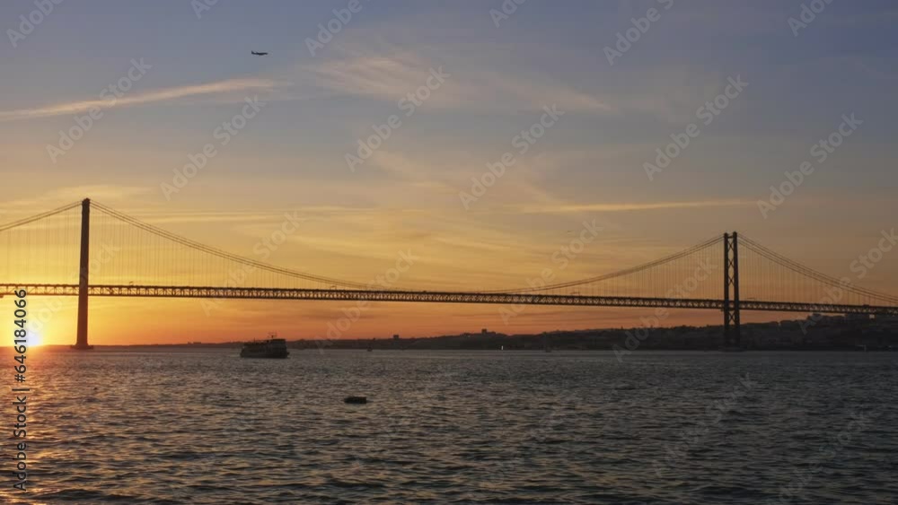 View of 25 de Abril Bridge famous tourist landmark of Lisbon connecting Lisboa and Almada on Setubal Peninsula over Tagus river with boat silhouette on sunset and flying plane. Lisbon, Portugal View of 25 de Abril Bridge famous tourist landmark of Lisbon connecting Lisboa and Almada on Setubal Peninsula over Tagus river with boat silhouette on sunset and flying plane. Lisbon, Portugal
