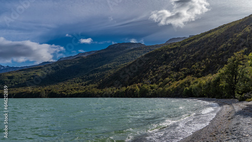 The beautiful emerald lake is surrounded by mountains. Green forest on the slopes. The waves are foaming on the pebbly shore. Clouds in the blue sky. Lago Roca. Tierra del Fuego National Park.