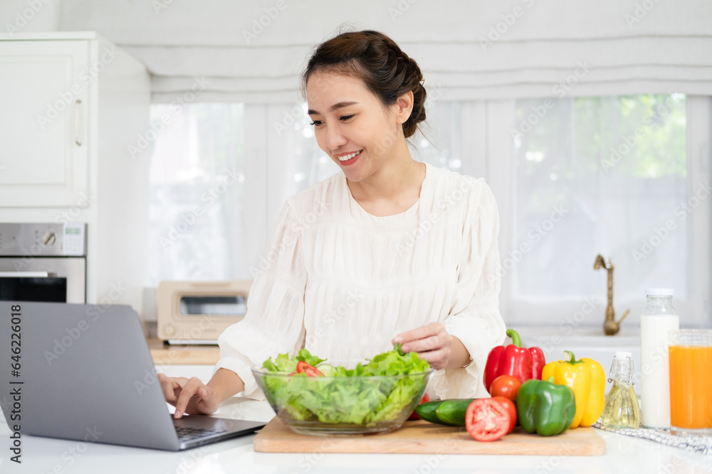 Online cooking tutorial. Asian young woman teenage girl using laptop for vlogging blogging looking for recipe while cooking vegetable vegan salad meal, healthy eating habits.