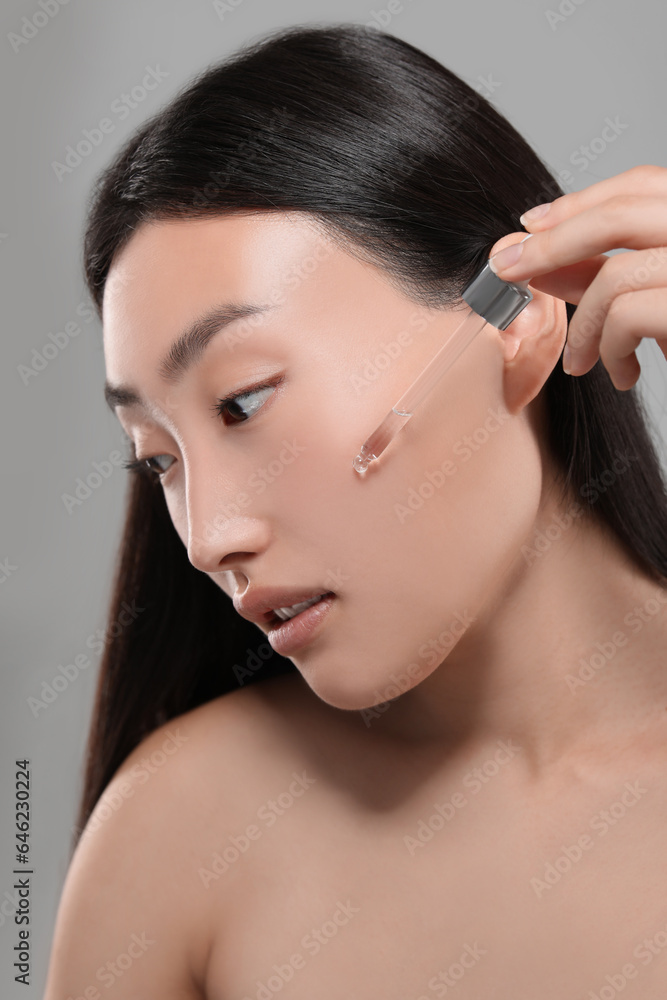 Beautiful young woman applying cosmetic serum onto her face on grey background
