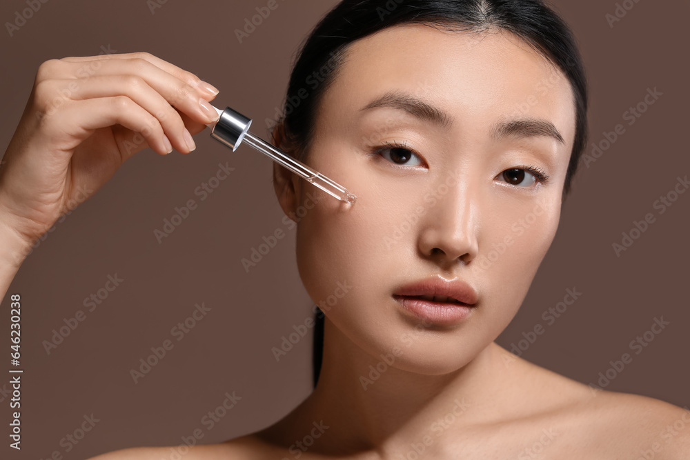 Fototapeta premium Beautiful young woman applying cosmetic serum onto her face on brown background, closeup