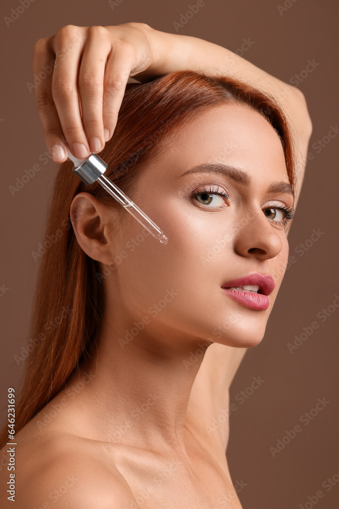 Beautiful young woman applying cosmetic serum onto her face on brown background