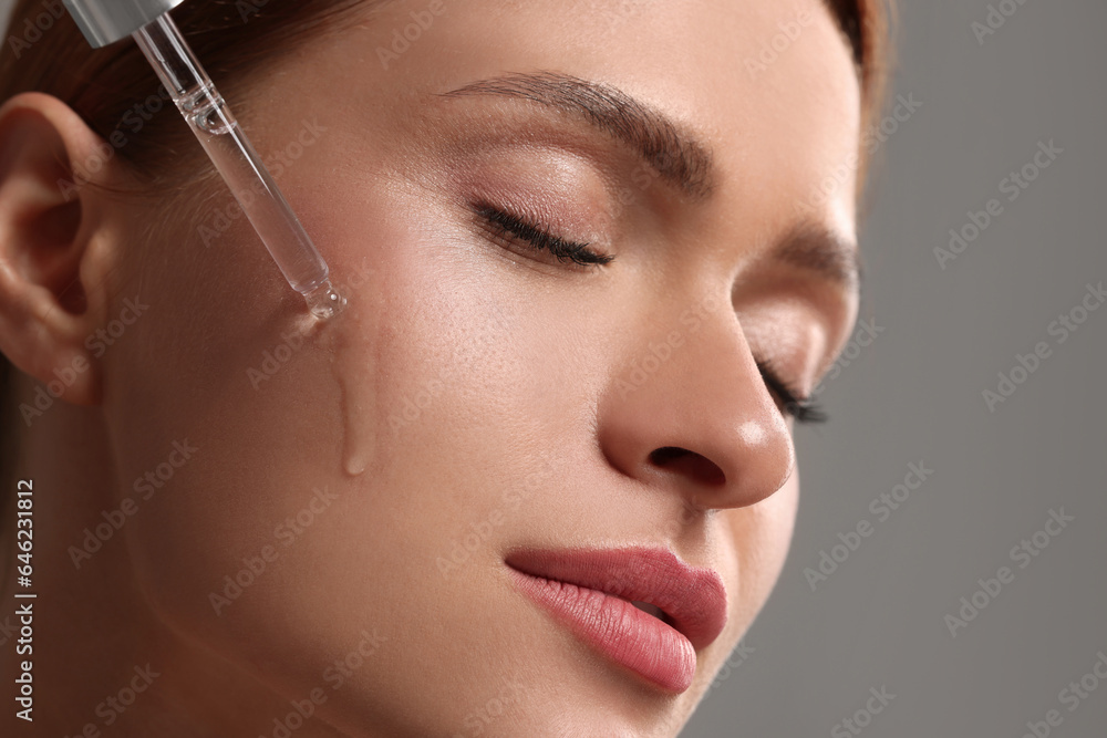 Beautiful young woman applying cosmetic serum onto her face on grey background, closeup