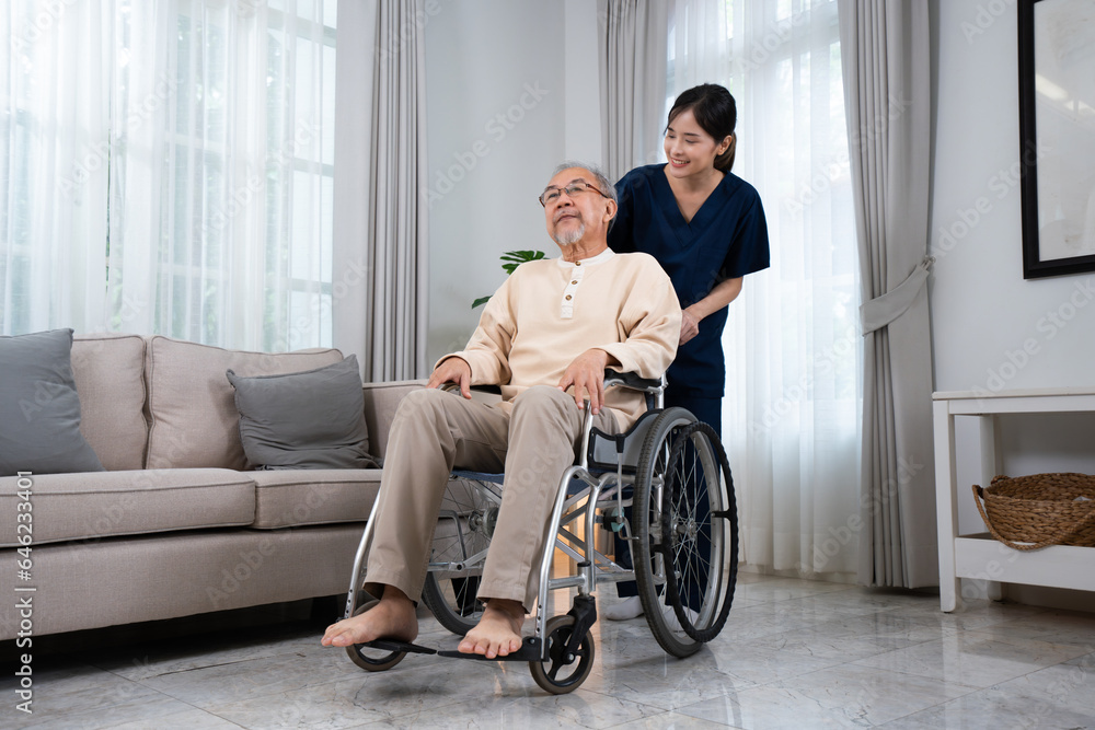 A young positive and happy Asian female nurse or physical therapist in a blue uniform, smiling pushing a senior Asian man sitting on a wheelchair while taking him to relax in the living room at home. A young positive and happy Asian female nurse or physical therapist in a blue uniform, smiling pushing a senior Asian man sitting on a wheelchair while taking him to relax in the living room at home.
